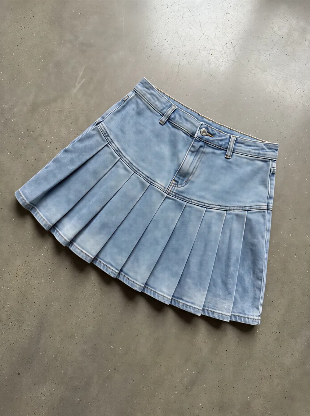 Product photo: a light wash pleated denim mini skirt laid flat on concrete